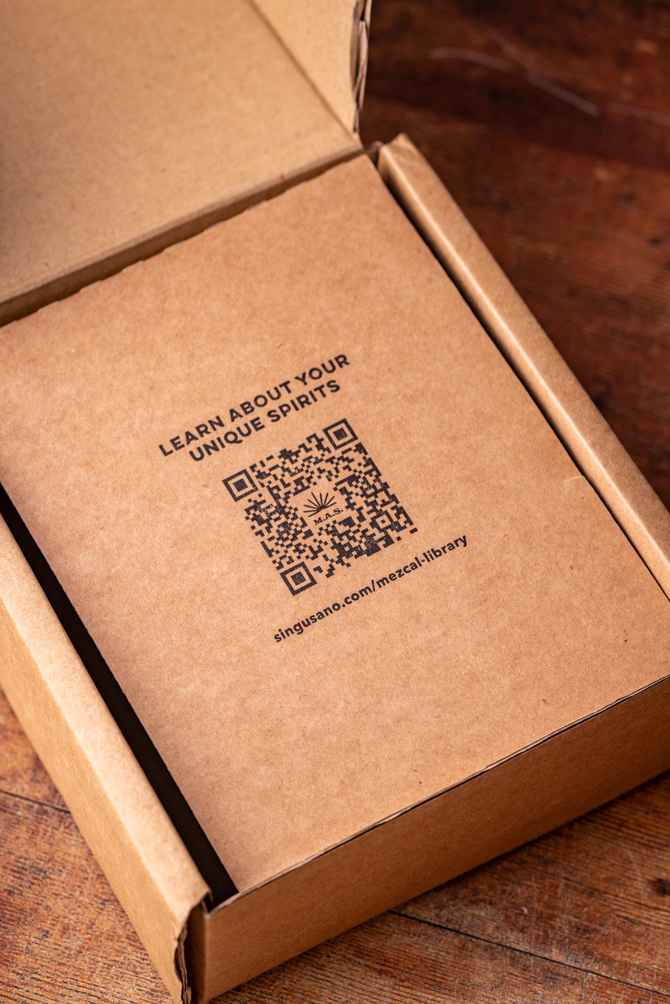 Brown cardboard box with a QR code linking to the Mezcal Library and text on a wooden surface