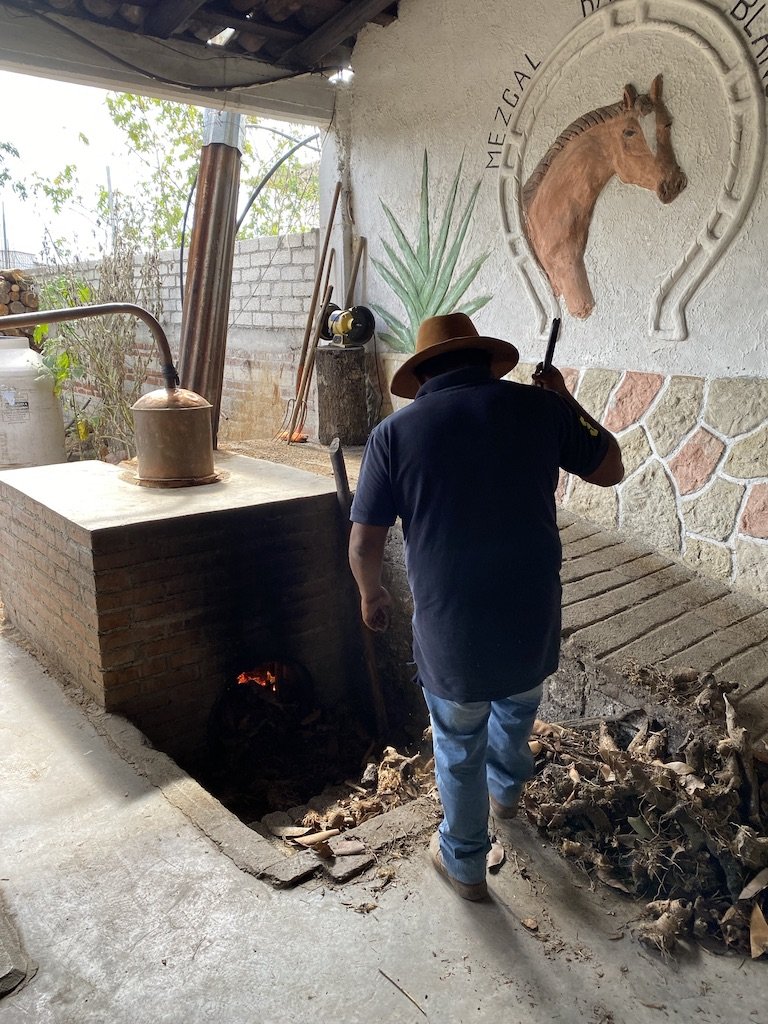 A man filling the fire below a copper pot still used to make mezcal