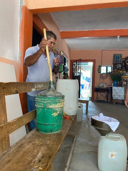 Nicolas, creator of the Tobala - Amatlán using a green funnel to transfer mezcal from a large container into a smaller one in a room.