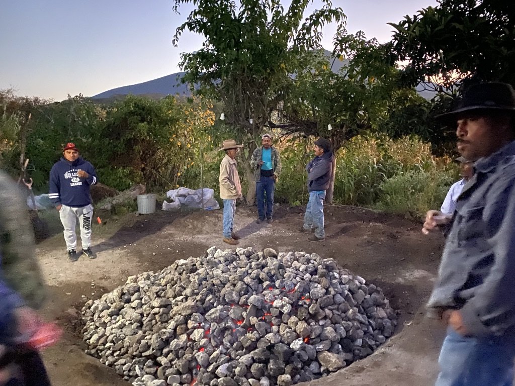 Group of people standing around an outdoor oven of rocks in an outdoor setting with trees and mountains in the background.