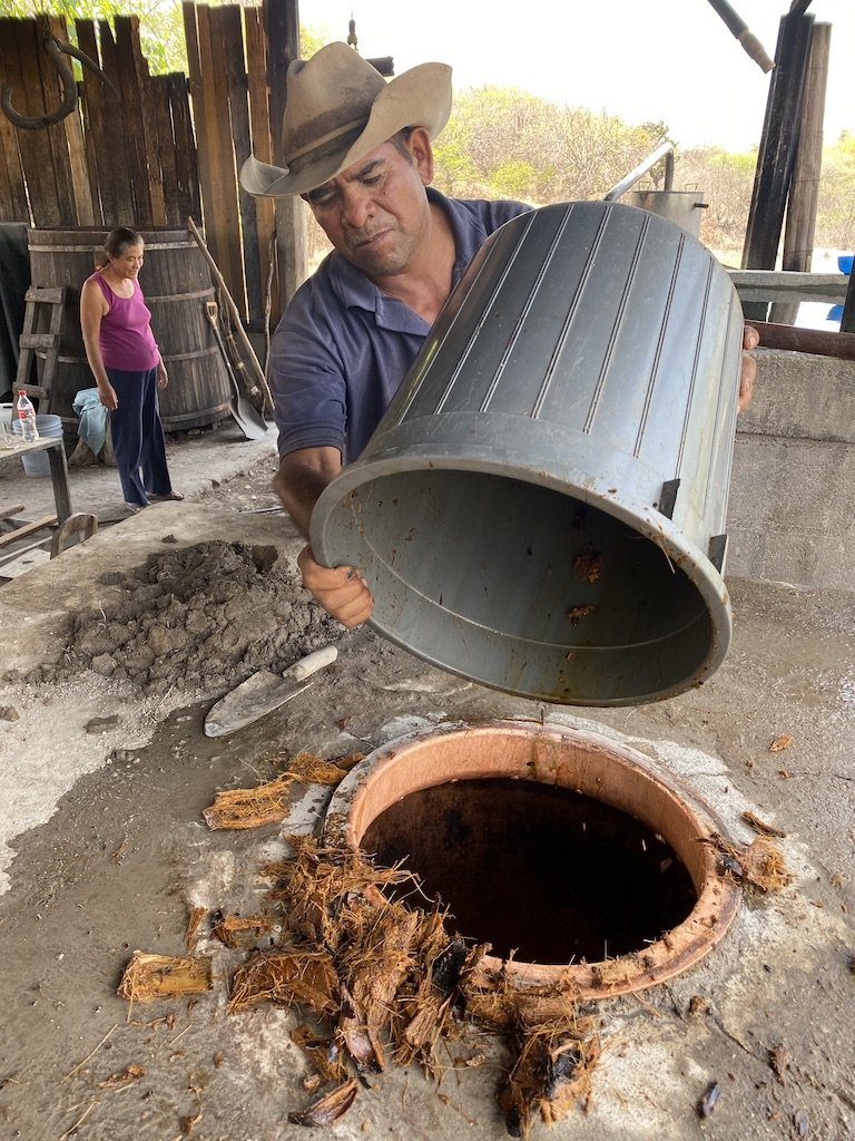 Man in a cowboy hat using a large metal container to handle all the fibre goes in the still for the first pass