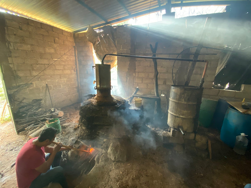 a small distillery with pot stills being heated by a man blowing on the embers through a tube in a shed in Mexico making mezcal