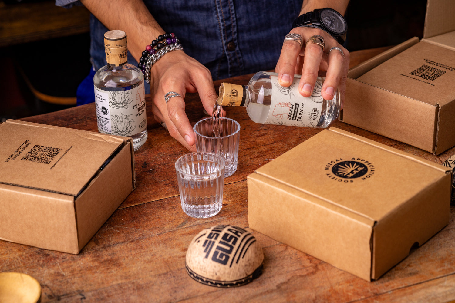 Person pouring mezcal from a bottle into small glasses on a wooden table with boxes from the Mezcal Appreciation Society and a bottle cap