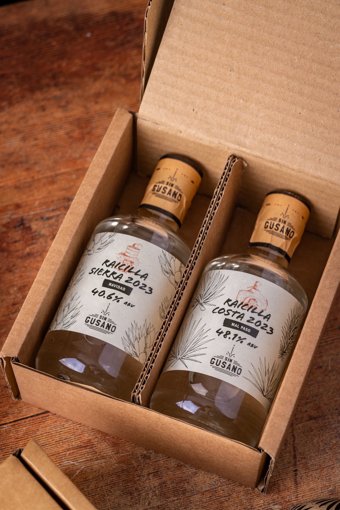 Two bottles of 'Cusano' mezcal on a wooden surface with a cardboard box.