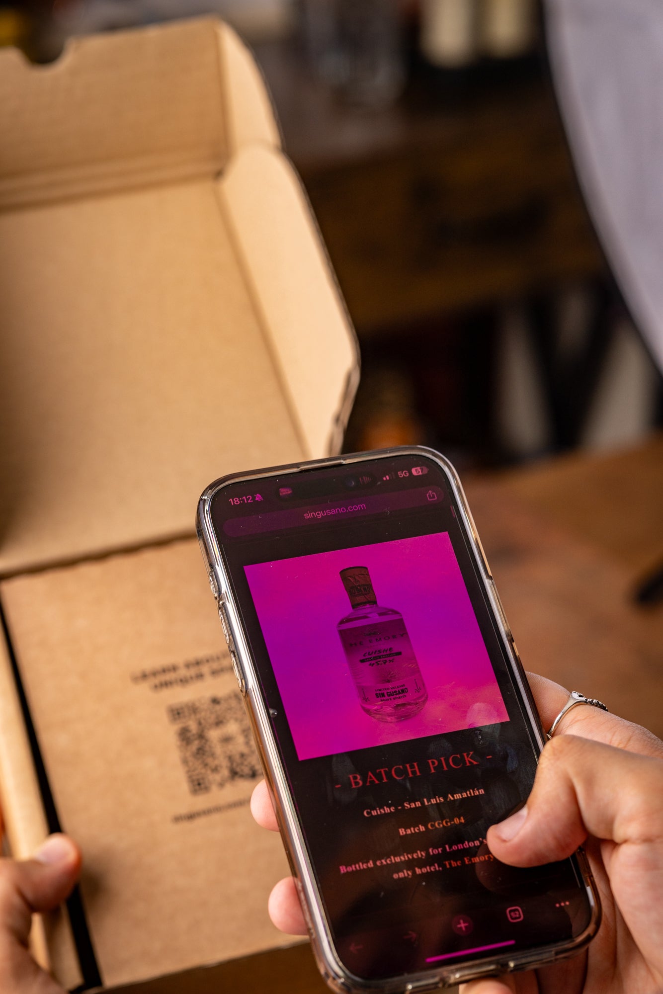 Person holding a phone with a bottle image on screen scanning a QR code on a cardboard box from the Mezcal Appreciation Society