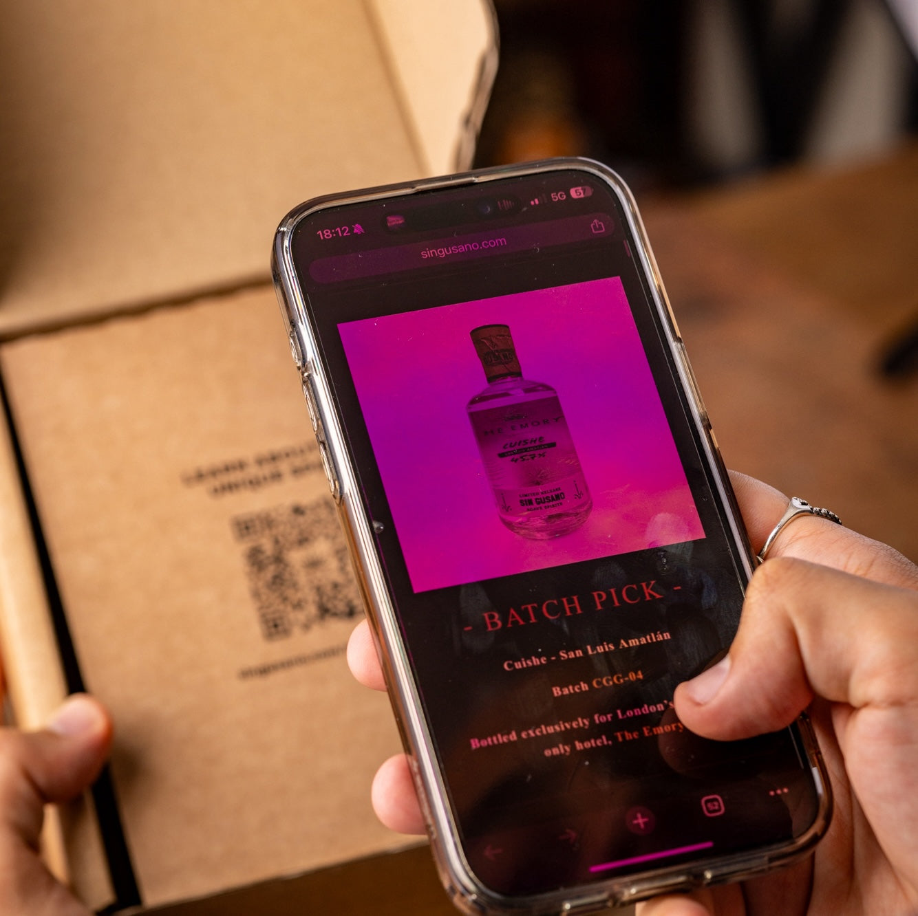 Person holding a phone with a bottle image on screen scanning a QR code on a cardboard box from the Mezcal Appreciation Society