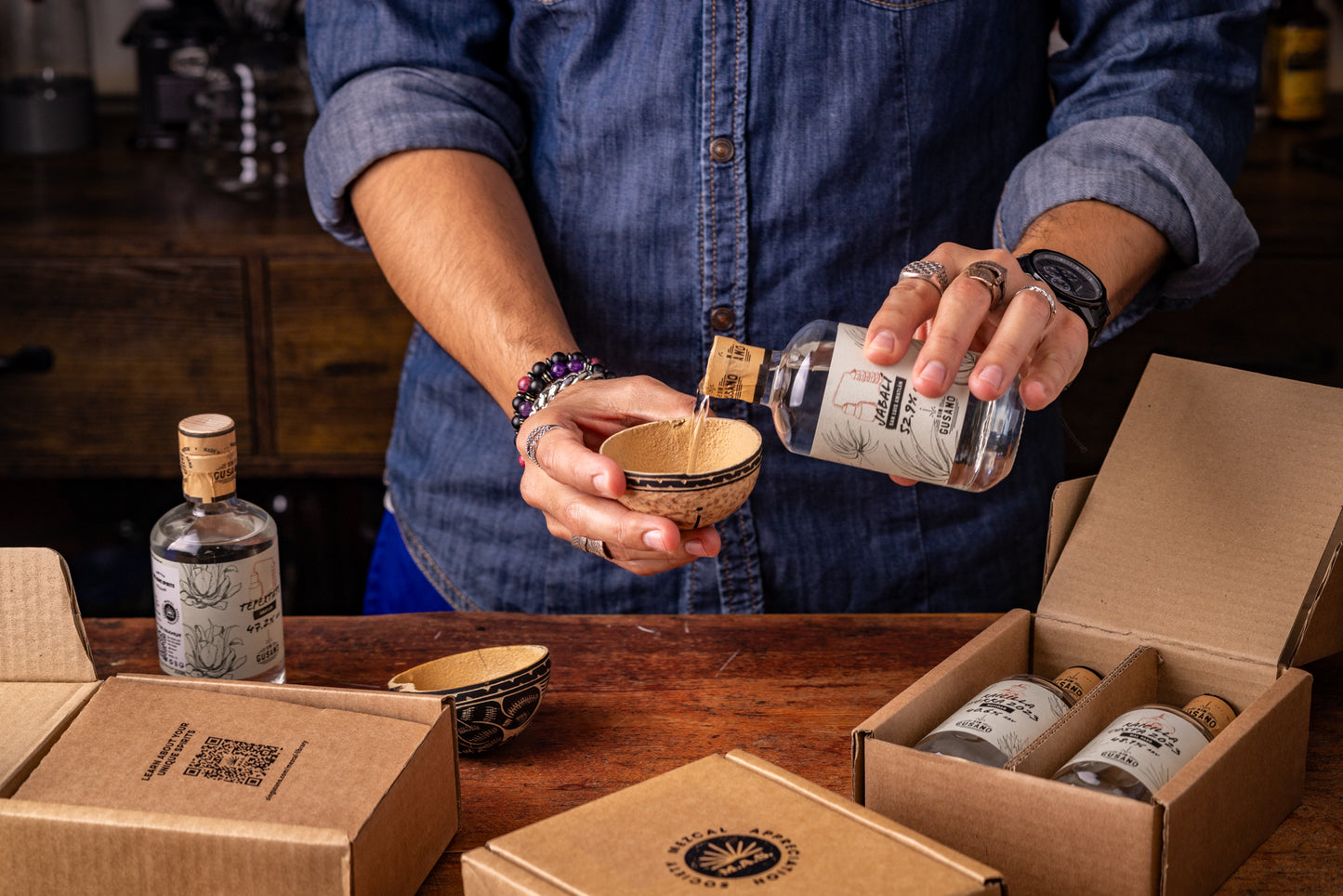 Person pouring mezcal from a bottle into a small bowl with boxes and bottles from the Mezcal Appreciation Society on a table.