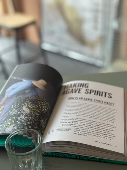 Open agave spirits book on a table with a glass of mezcal, blurred background
